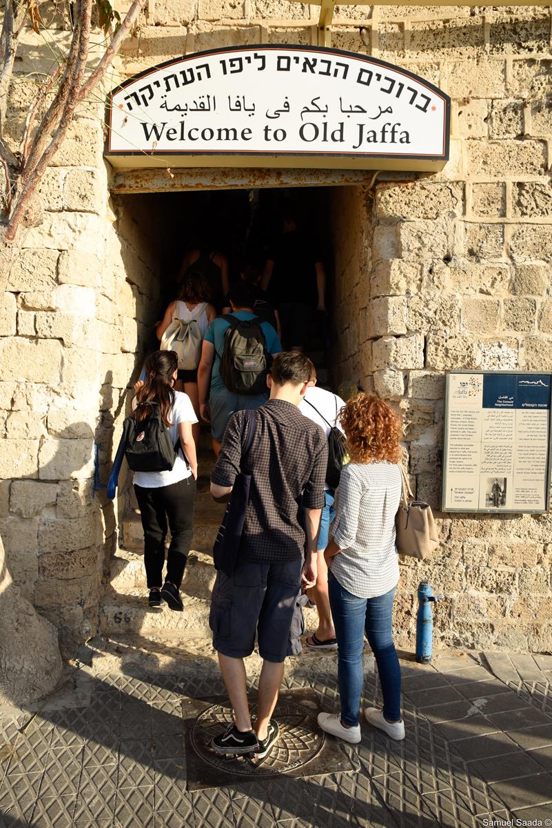 Uczestnicy programu podczas spaceru z przewodnikiem po starej Jafie, wchodzą w bramę znajdującą się w murze zbudowanym z kamienia w kolorze piasku