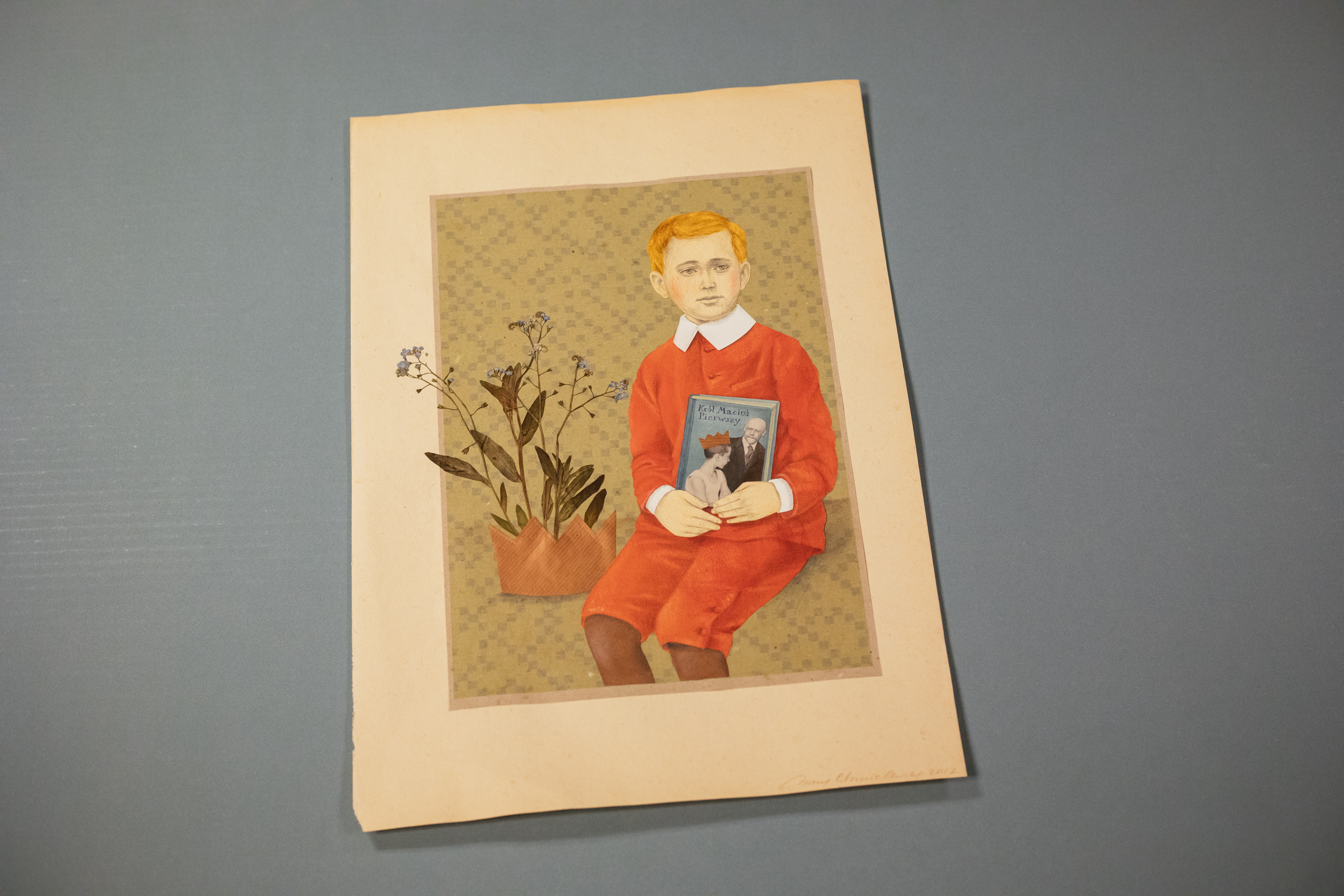 Obraz przedstawiający króla Maciusia -rudego chłopca w czerwonej marynarce i czerwonych spodniach do połowy nóg, trzymającego przy pasie, okładką do widza, książkę. Na lewo od chłopca - niebieskie kwiatki, wystające z brązowej schematycznej korony.