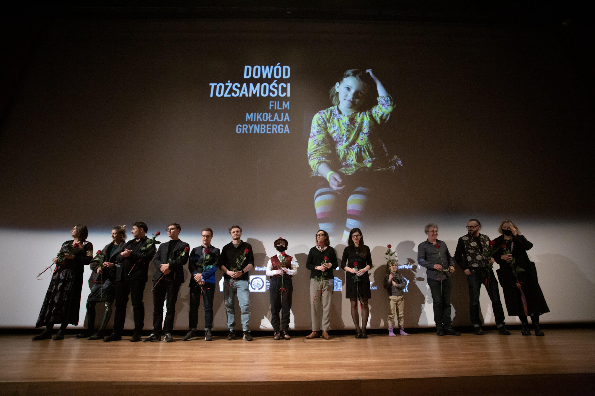 Na scenie w rzędzie stoi grupa ludzi. W tle bohaterka filmu "Dowód tożsamości", Chana Topińska oraz napis z tytułem filmu i podtytułem: Film Mikołaja Grynberga.