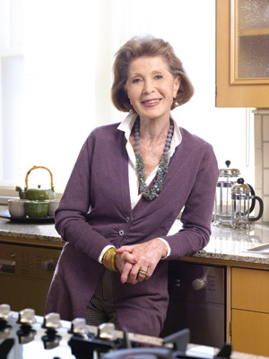 Helen Lash. Older lady. She wears violet jacket and grey necklace. She is in the kitchen.