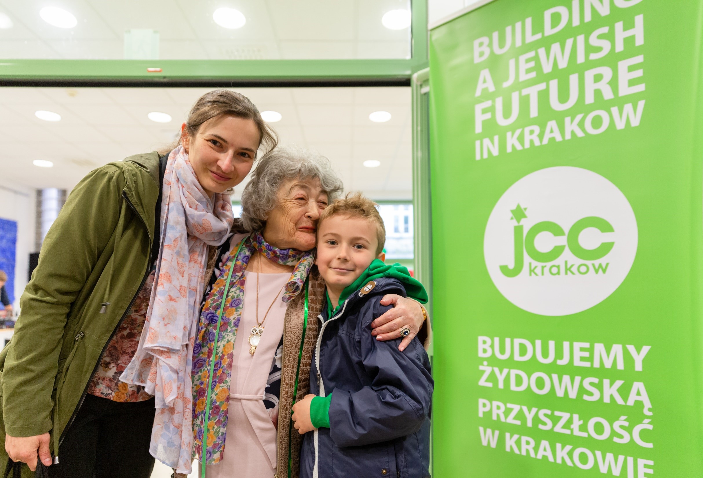 Dwa pokolenia kobiet - młoda i starsza - i chłopiec ściskają się. Obok baner z logo JCC Kraków. Budujemy żydowską przyszłość w Krakowie. 