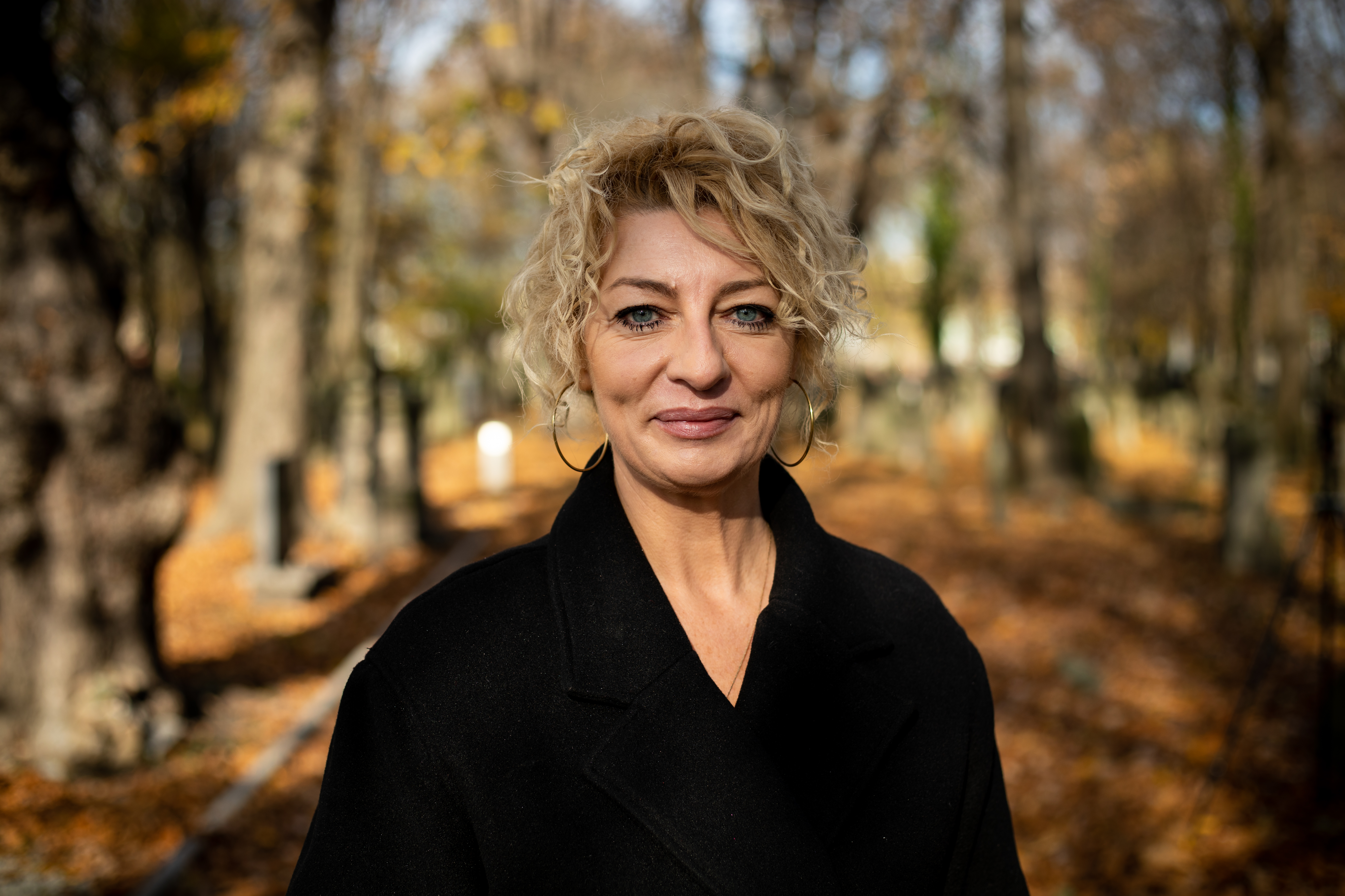 Inga Marczyńska w czarnym płaszczu, stoi w otoczeniu drzew na cmentarzu żydowskim. Jest piękna złota jesień.