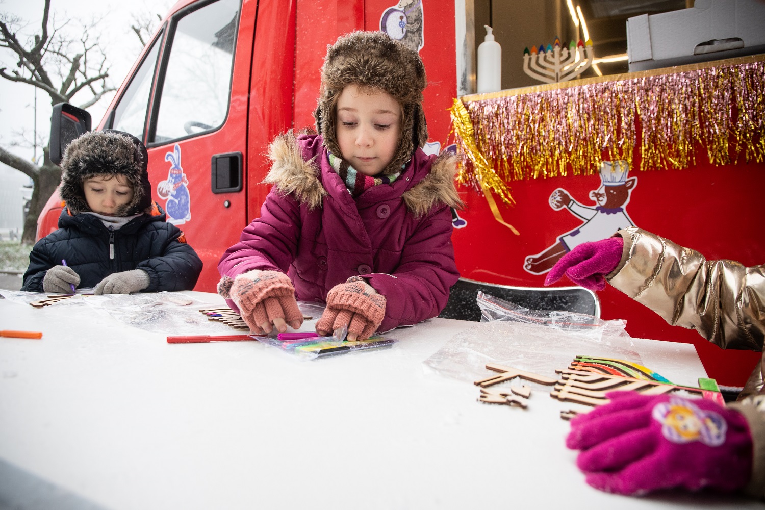 Dzieci podczas warsztatów w trakcie Rodzinnej Chanuki 2021 ozdabiają modele chanukij (świec chanukowych). W tle czerwony chanukobus.