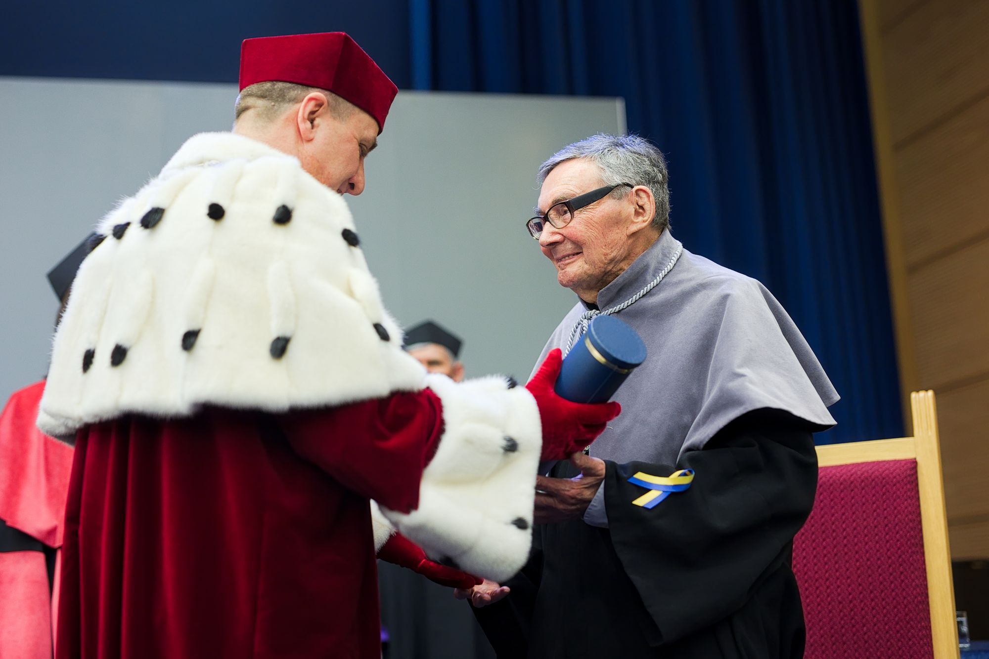 Marian Turski odbiera z rąk rektora UMCS tytuł honoris causa. Rektor nosi czerwoną czapkę i togę, a na plecach ma białe futerko. Marian Turski ma czarną szatę i szarą pelerynkę.