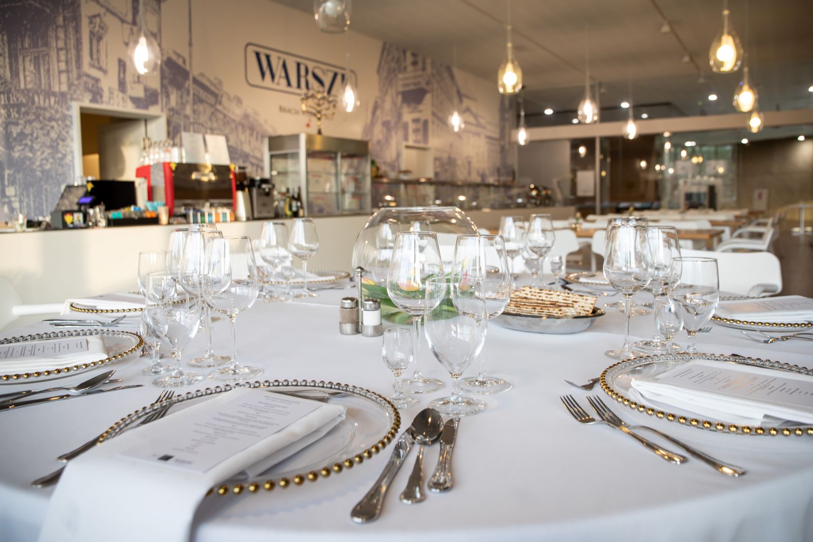 Biały stół ze sztućcami, kieliszkami i talerzami. W tle logo restauracji "Warsze".