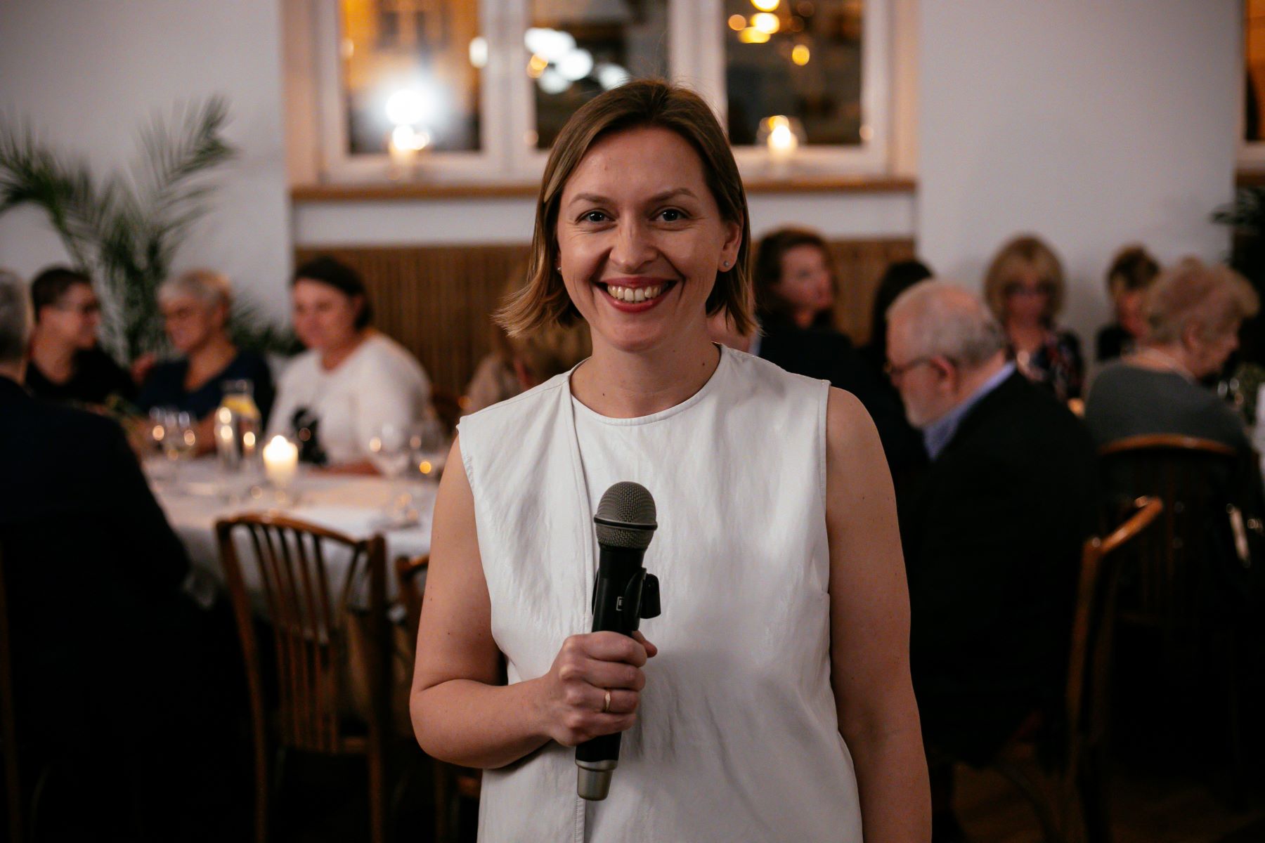 Uśmiechnięta Magdalena Maślak stoi z mikrofonem. Za nią przy stole siedzi kilkanaście osób.