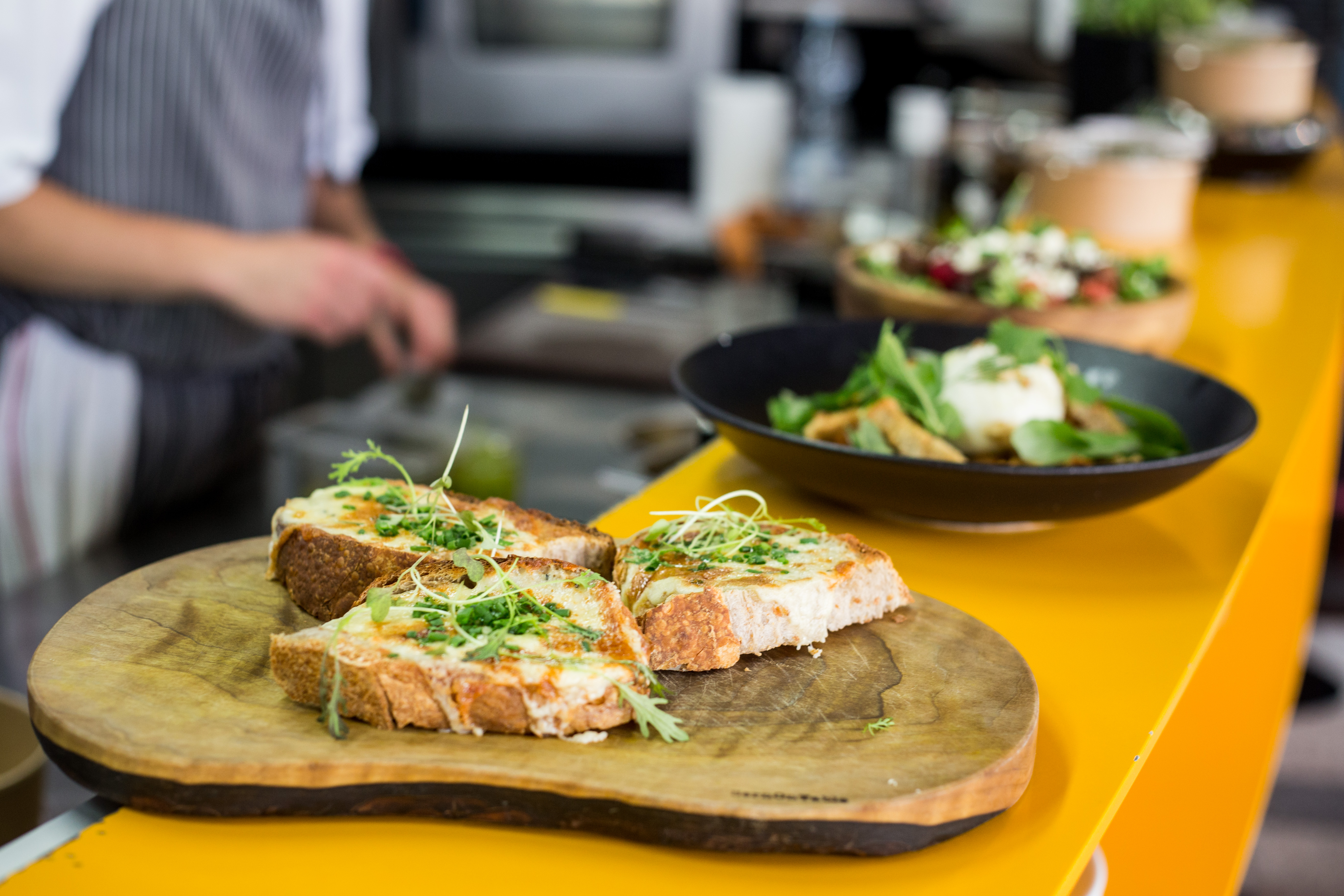 Na drewnianej desce, na barze w restauracji, leżą trzy grillowane kanapki z pesto. W tle widać inne dania w talerzach i kucharza, który przygotowuje inne danie.