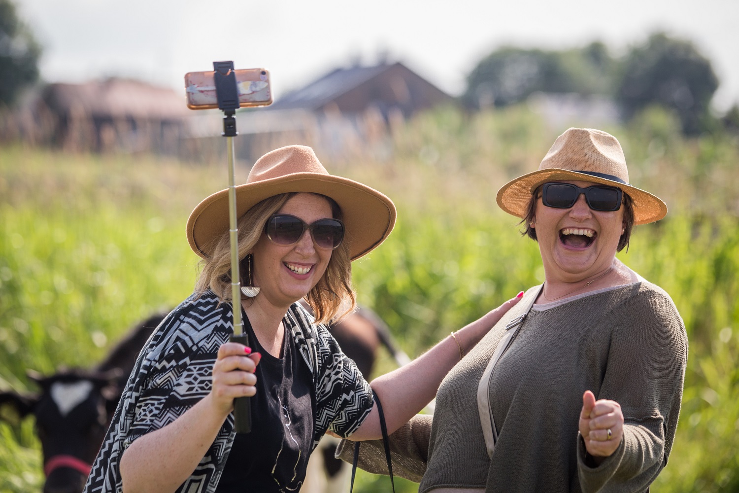 Dwie kobiety w ciemnych okularach i jasnych kapeluszach, robią selfie. Bardzo roześmiane. W tle łąka i zabudowania wiejskie.