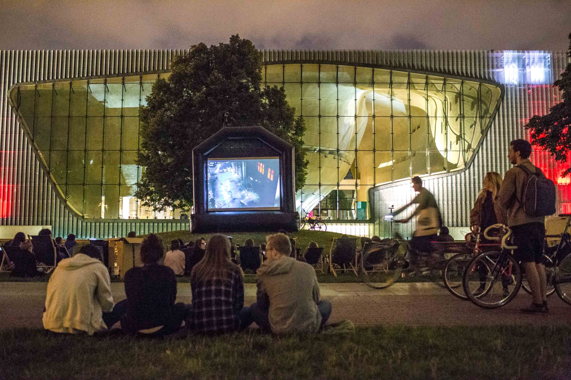 Widzowie siedzą na trawie i oglądają film wyświetlany na ekranie podczas pokazu kina letniego POLIN.