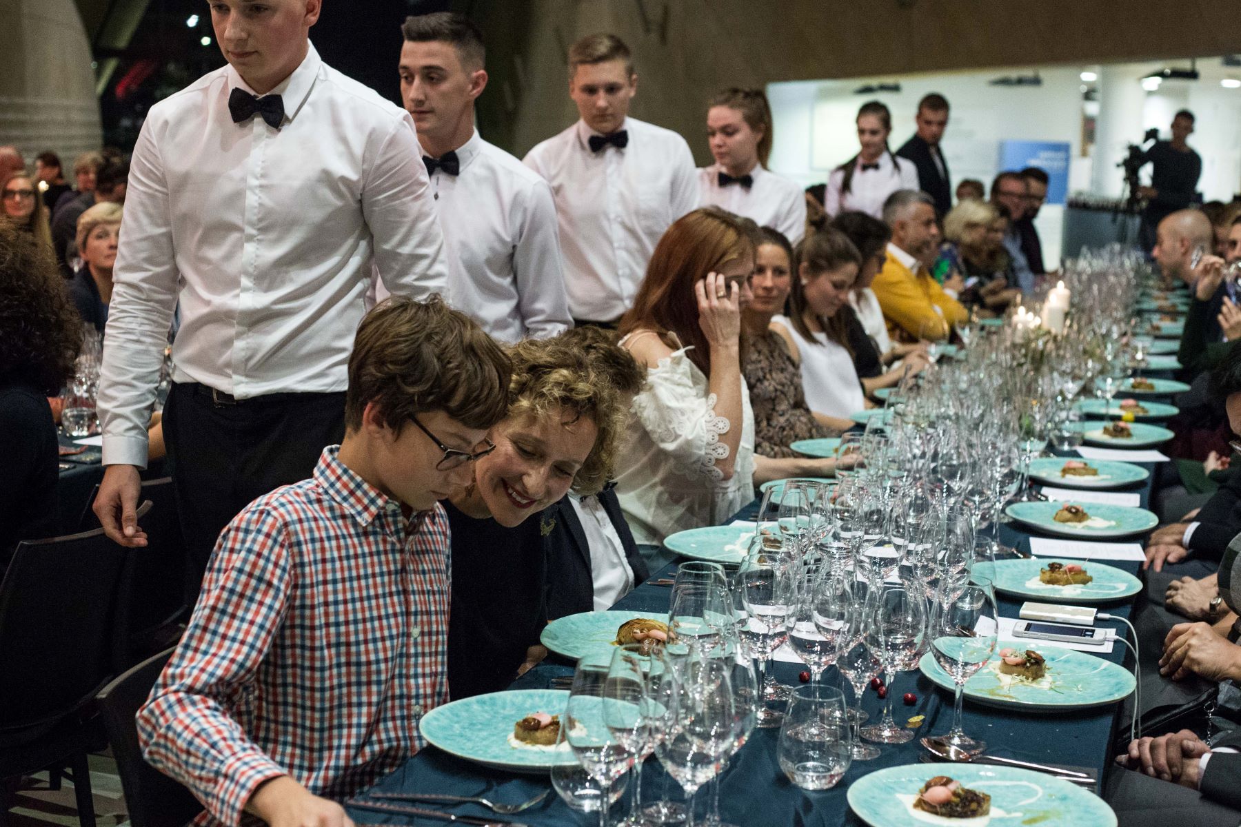 Grupa uczestników kolacji szabatowej siedzi przy zastawionym stole. Wokół kelnerzy w koszulach i muszkach.