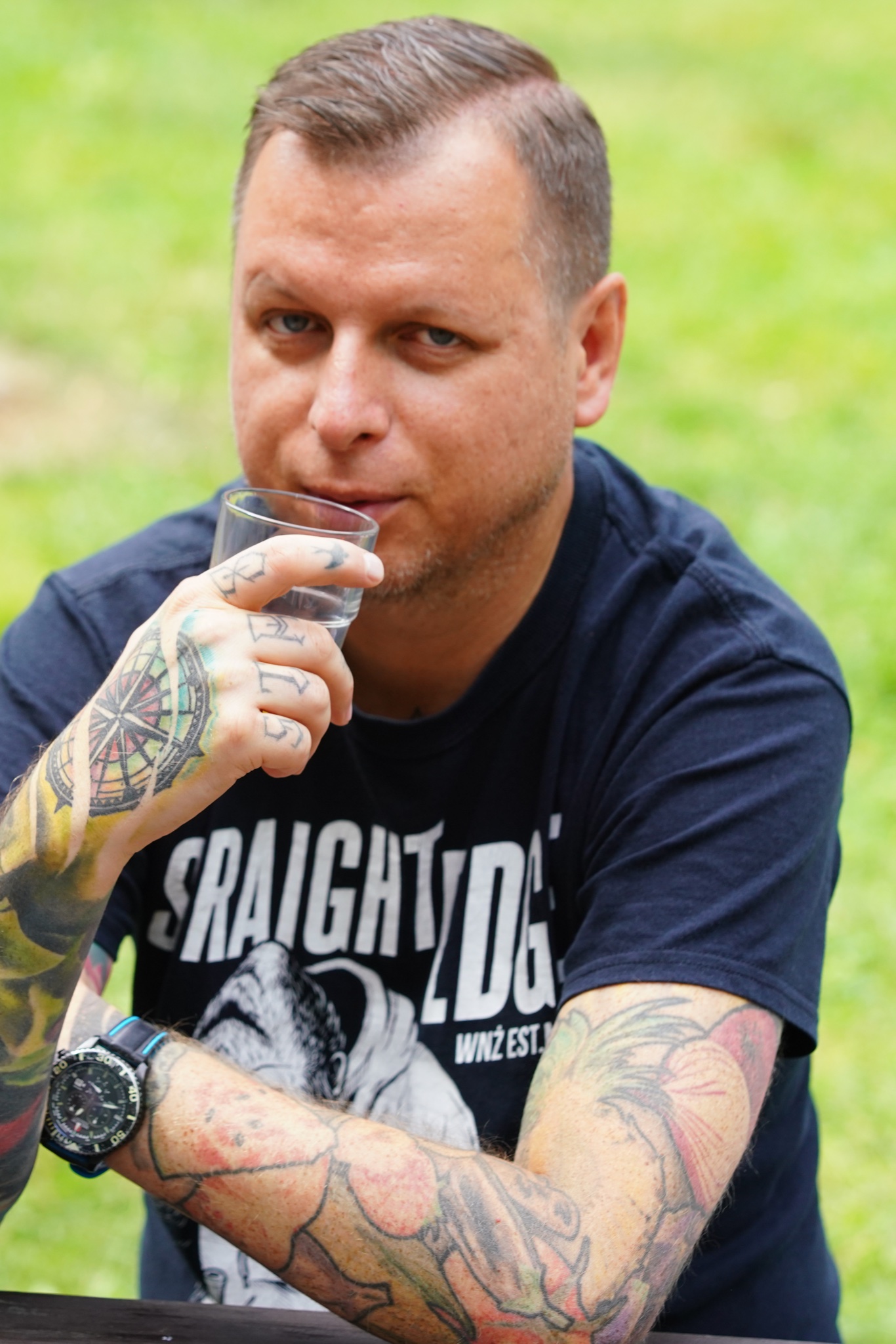 Daniel Jaroć trzyma szklankę przy twarzy. Na rękach i dłoniach ma tatuaże.