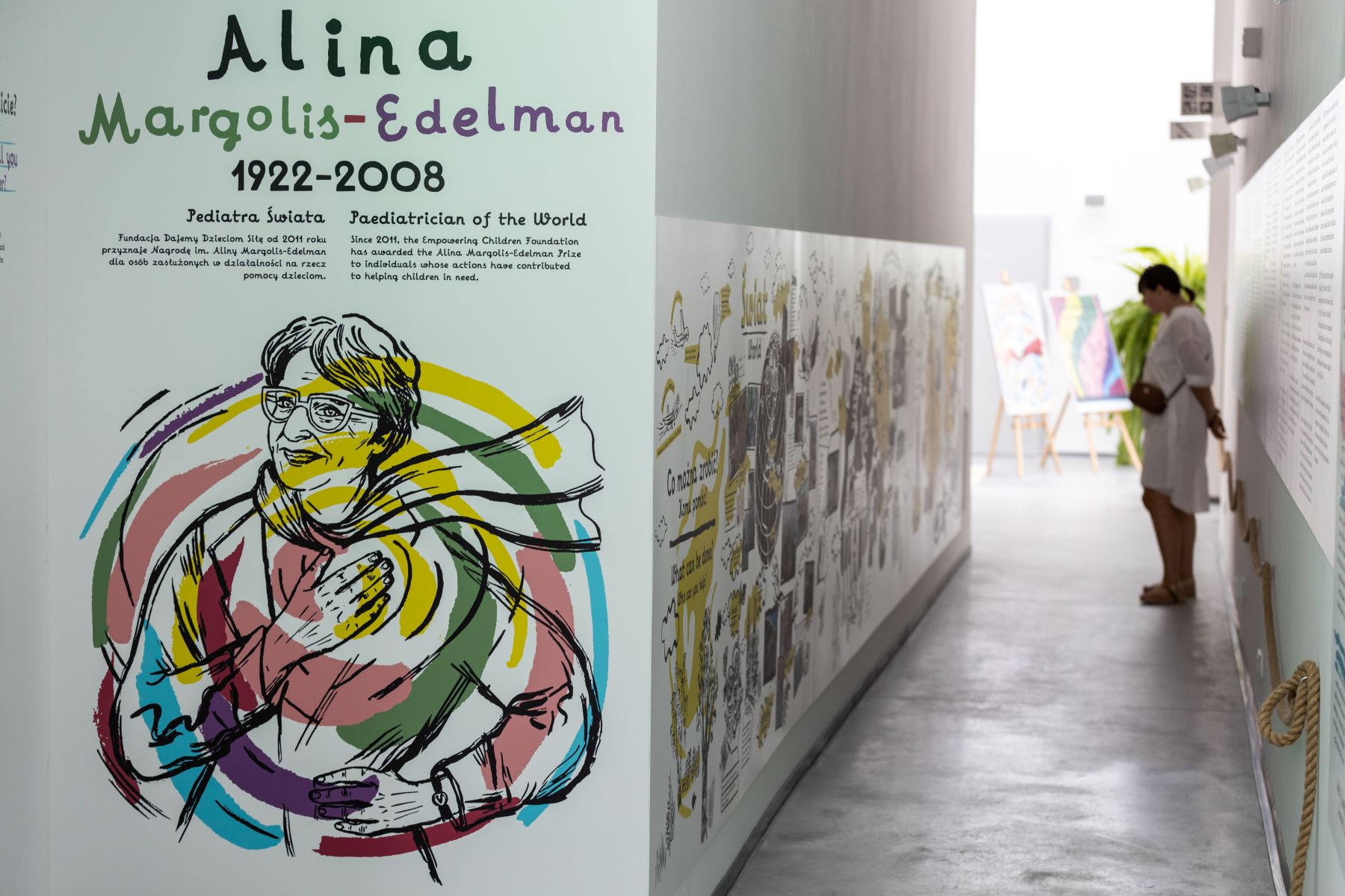 Wystawa "Elementarz empatii" w Łodzi. Zwiedzająca ogląda obrazki narysowane na ścianie. Przy wejściu na ekspozycję kolorowy rysunek przedstawiający postać Aliny Margolis-Edelman.