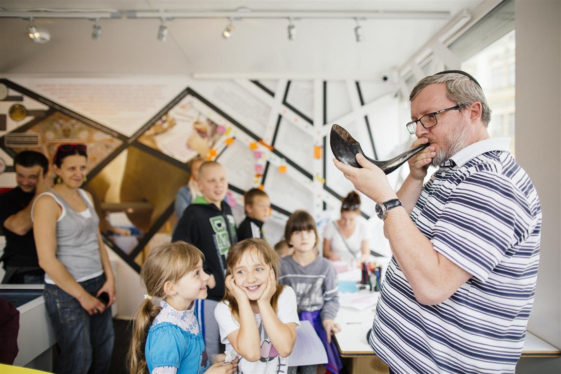 Zajęcia edukacyjne w pawilonie "Muzeum na kólkach". Mężczyzna w jarmułce prezentuje dzieciom dźwięk szofaru, czyli tradycyjnego instrumentu używanego w judaizmie.