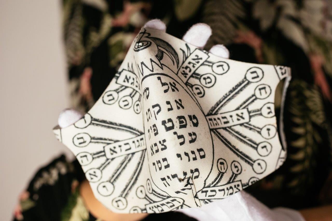 Ktoś trzyma przed sobą maseczkę z wzorami nawiązującymi do tych z żydowskiego amuletu ochronnego.
