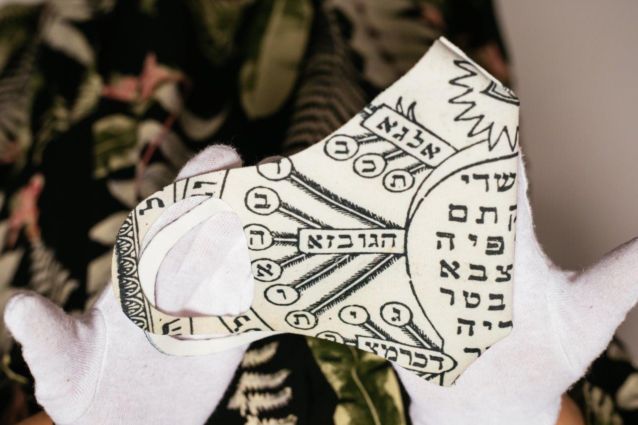 Ktoś trzyma przed sobą złożoną na pół maseczkę zdobioną inskrypcją w języku hebrajskim.