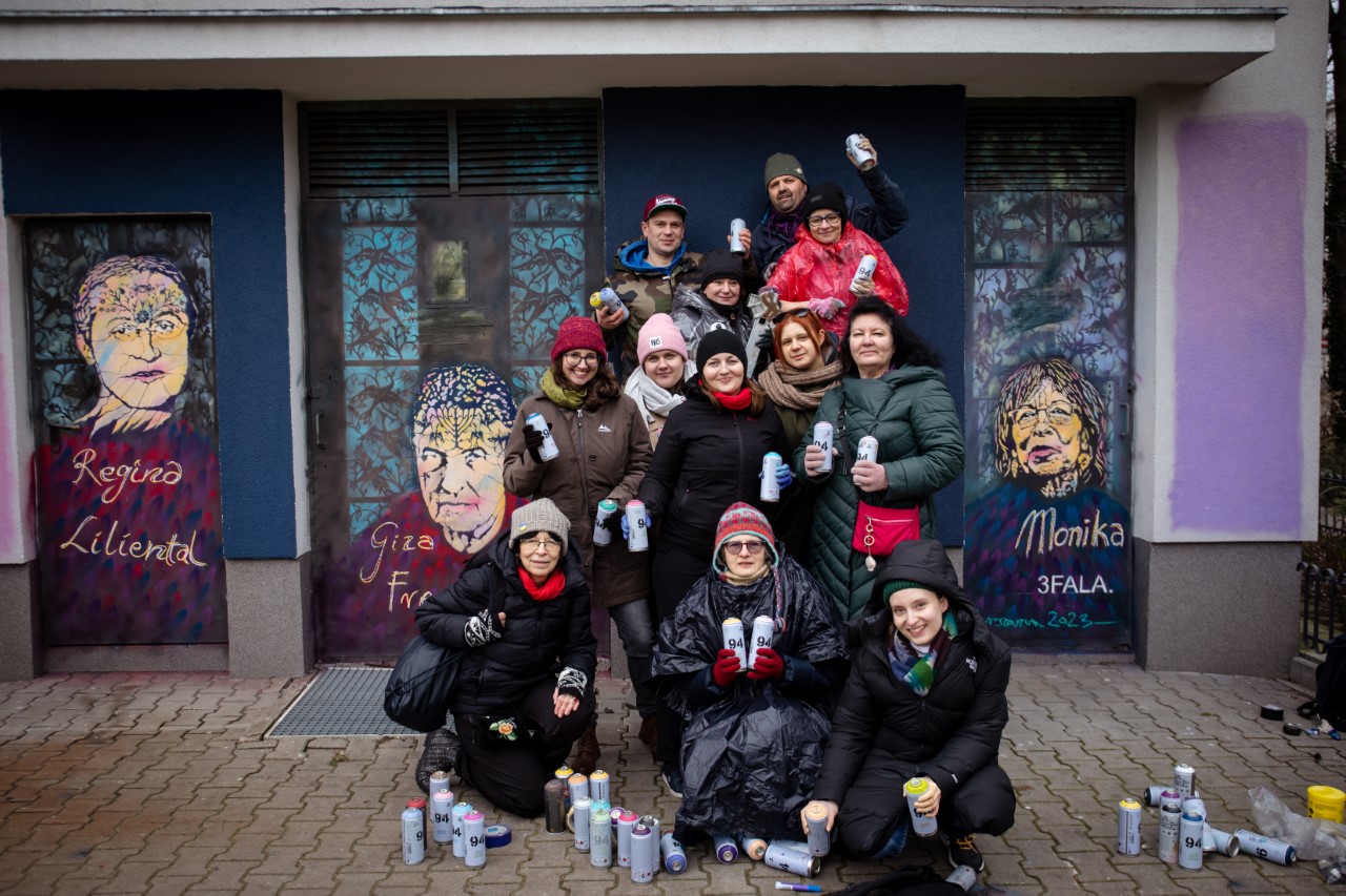 Grupa pracowników Muzeum POLIN trzyma w dłoniach spraye. Za nimi gotowy kolorowy mural.
