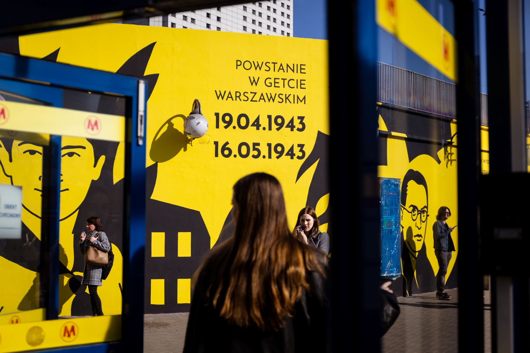 Dziewczyna stoi w drzwiach metra Centrum w Warszawie. W tle mural upamiętniający cywilów podczas powstania w getcie warszawskim.