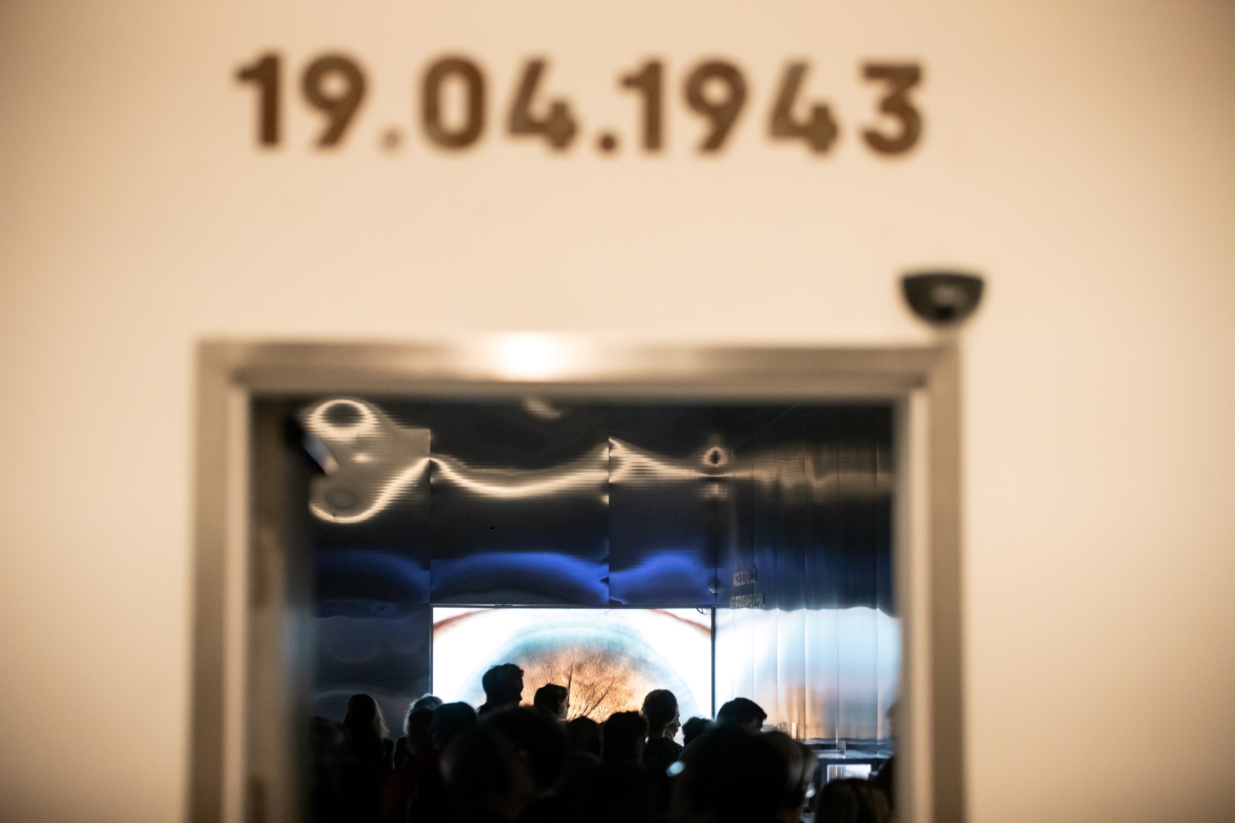 Wejście na wystawę "Wokół nas morze ognia". Nad nim data 19.04.1943. W tle zwiedzający podczas wernisażu.