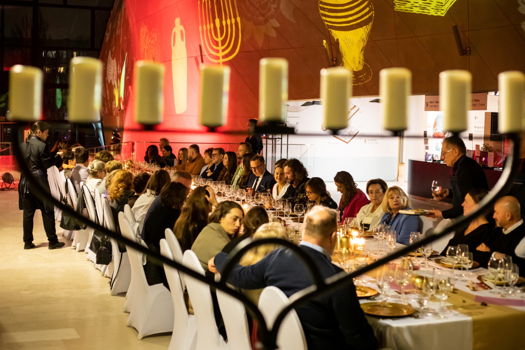Przy stole w holu głównym Muzeum POLIN siedzą goście kolacji szabatowej. Na ścianie wyświetlają się symbole związane z tradycją żydowską, np. świeca chanukija.