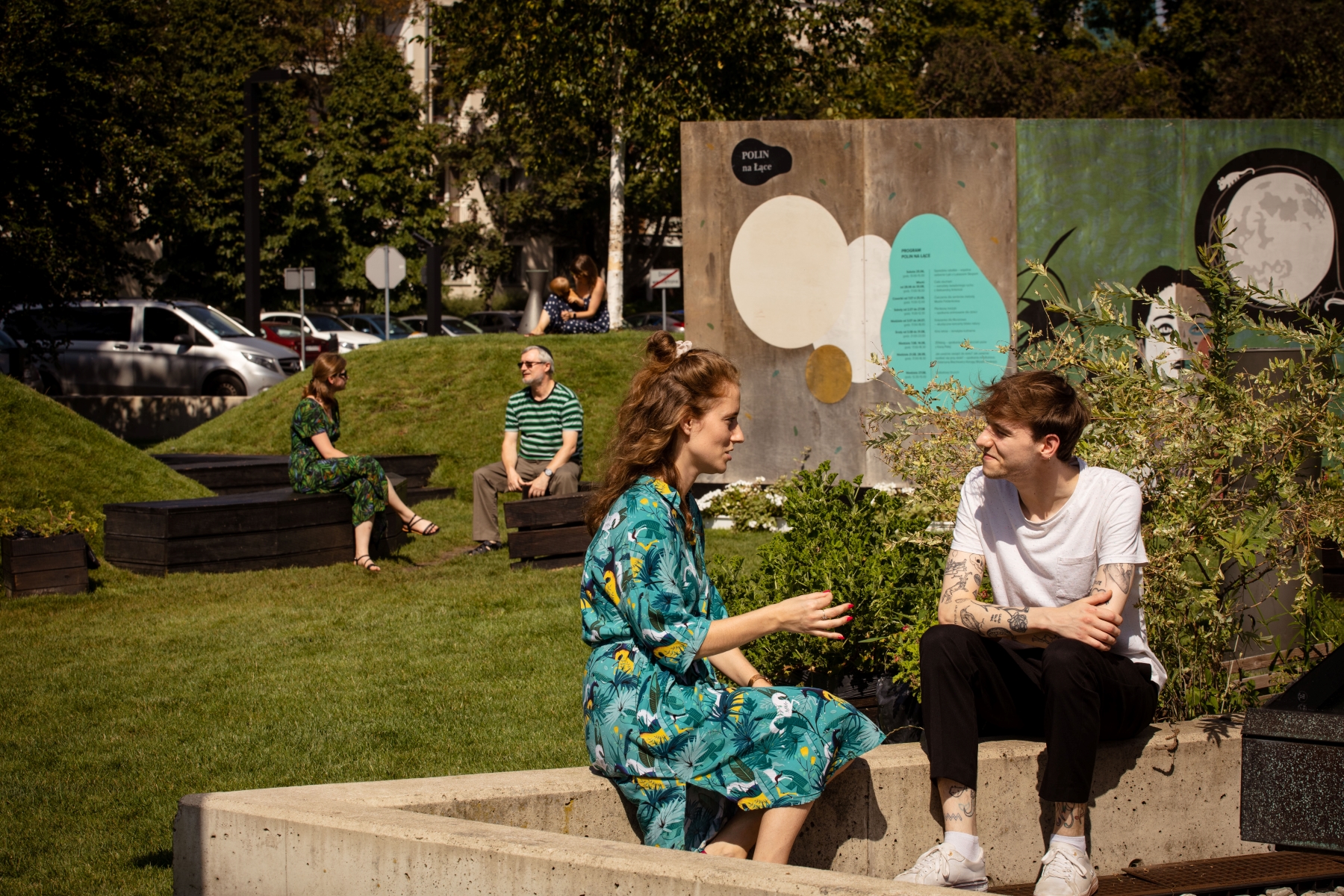 Dziewczyna i chłopak rozmawiają ze sobą, siedząc na murku przy instalacji "POLIN na Łące". W tle, przy muralu siedzą naprzeciw siebie dwie inne osoby.