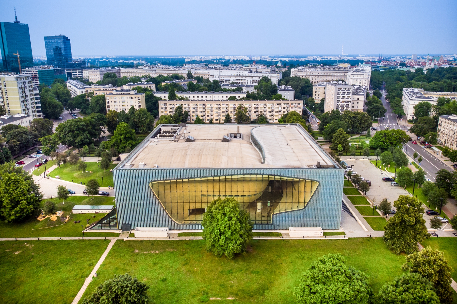Budynek Muzeum POLIN w otoczeniu drzew, zieleni, bloków na Muranowie i wieżowców Intraco przy Dworcu Gdańskim.