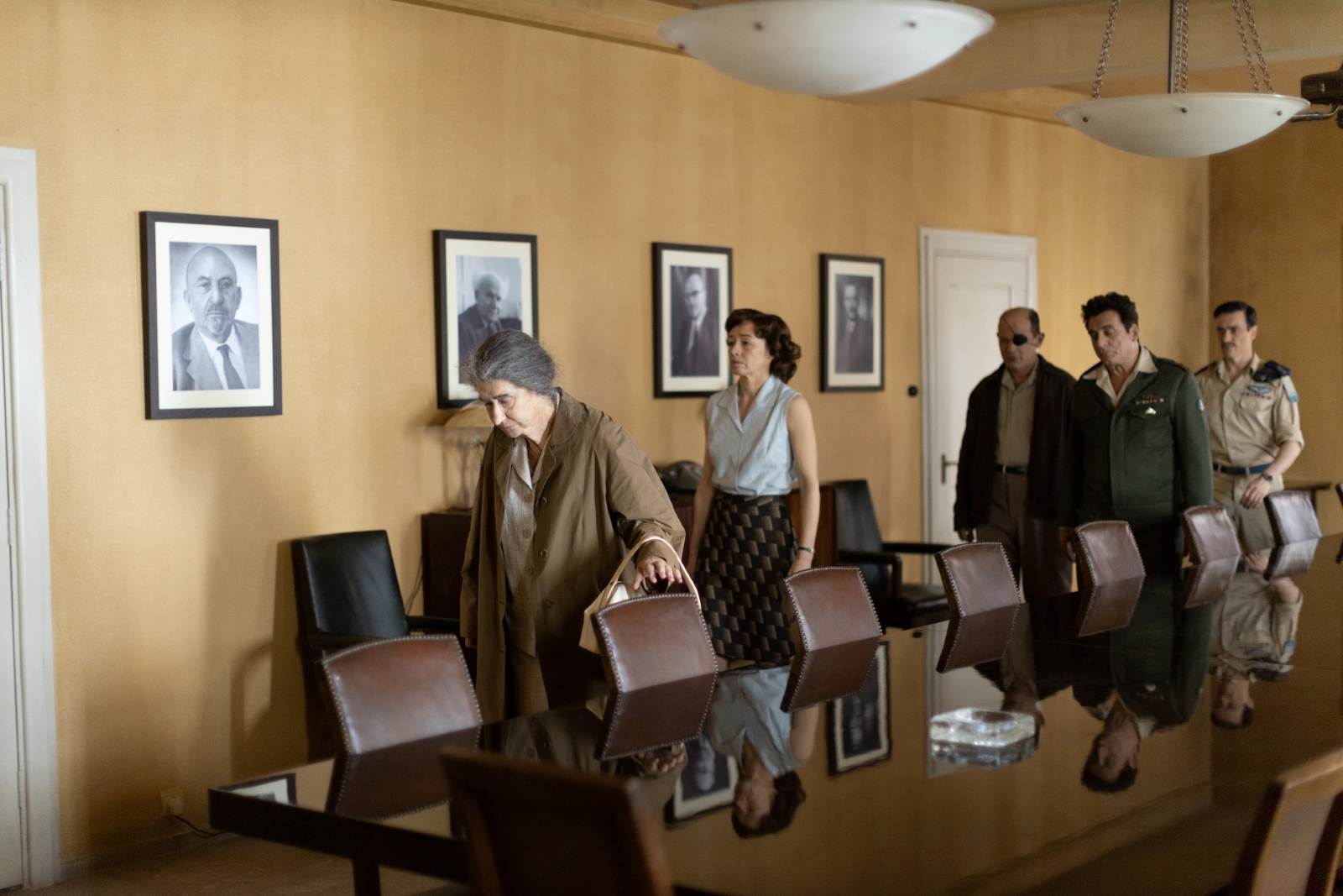 Kadr z filmu "Golda" - za główną bohaterką stoją kolejno: kobieta i trzej mężczyźni. Wszyscy są w jakimś pomieszczeniu i znajdują się za długim stołem.