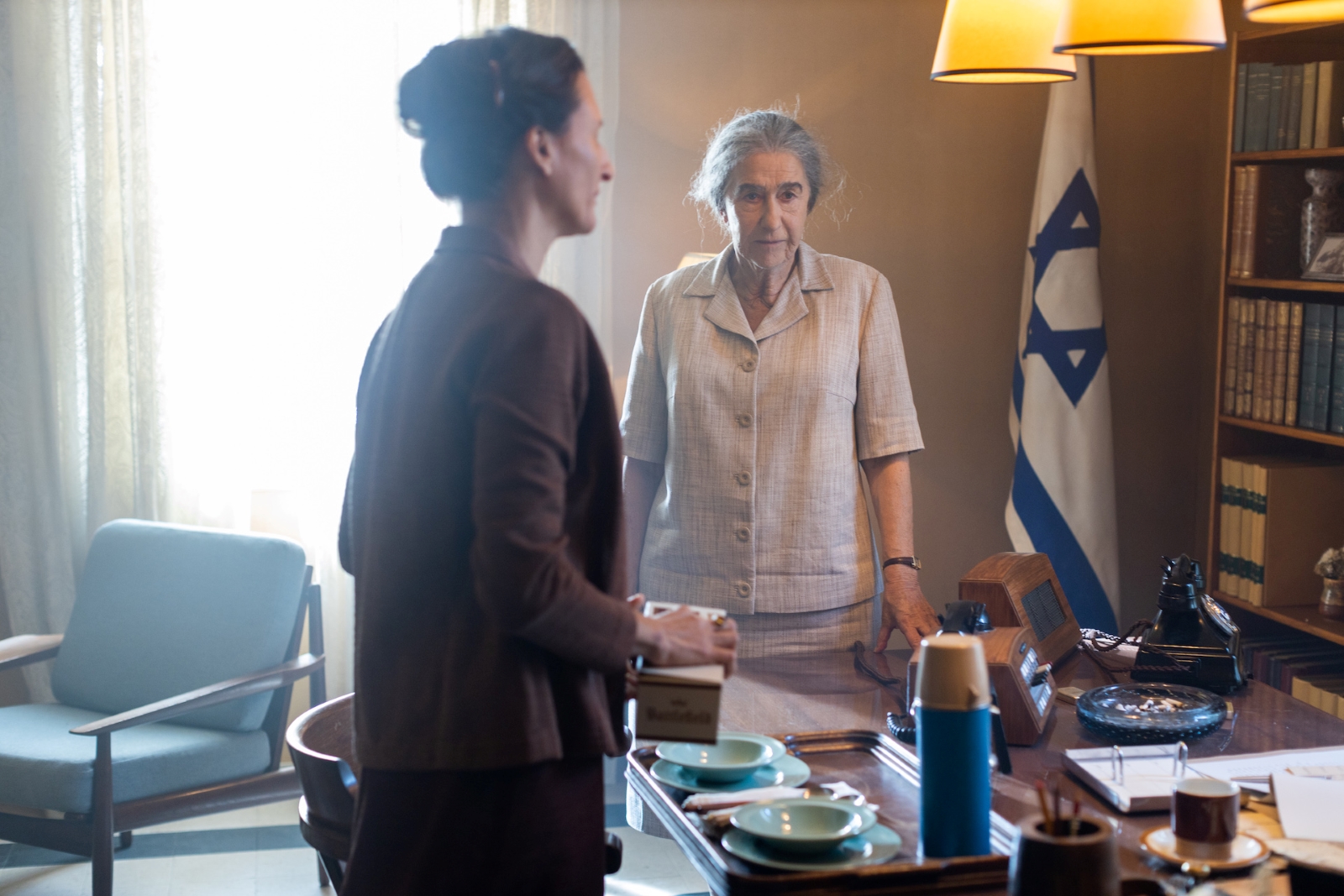 Kadr z filmu "Golda" - główna bohaterka stoi w gabinecie naprzeciwko innej kobiety. W tle flaga Izraela.