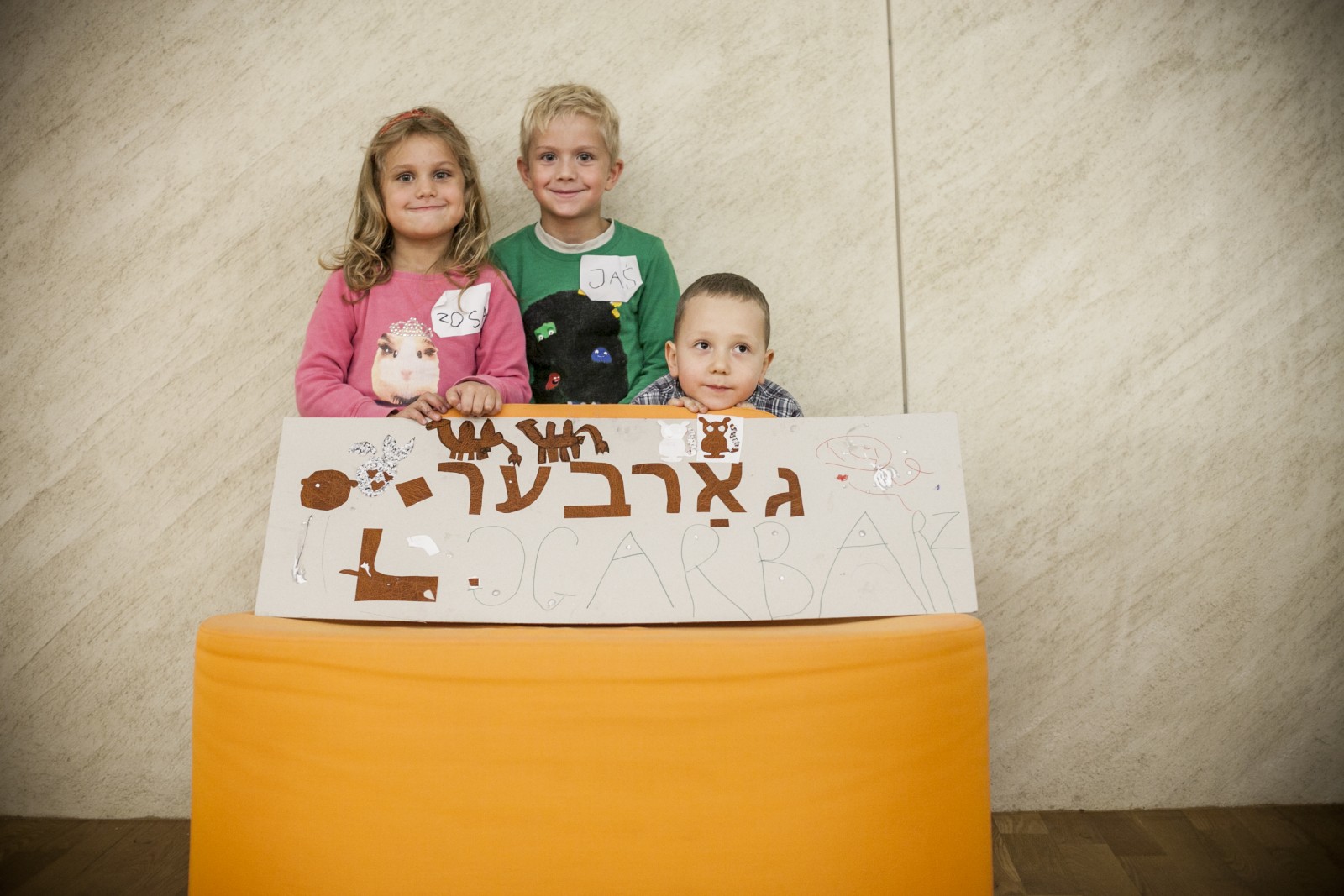 Troje dzieci stoi za pomarańczowym pufem, na którym stoi kartonowa tabliczka z literami hebrajskimi i polskimi. Wszyscy uśmiechają się radośnie, chłopiec z prawej strony jest schylony