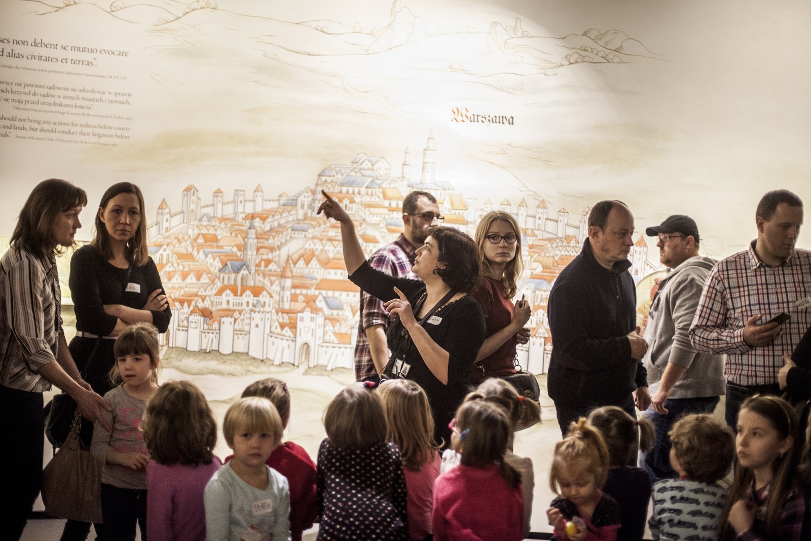 Grupa dzieci, za nimi grupa dorosłych, jedna z kobiet wskazuje coś wysoko w górze. Za plecami, w tle, średniowieczne przedstawienie planu Warszawy.