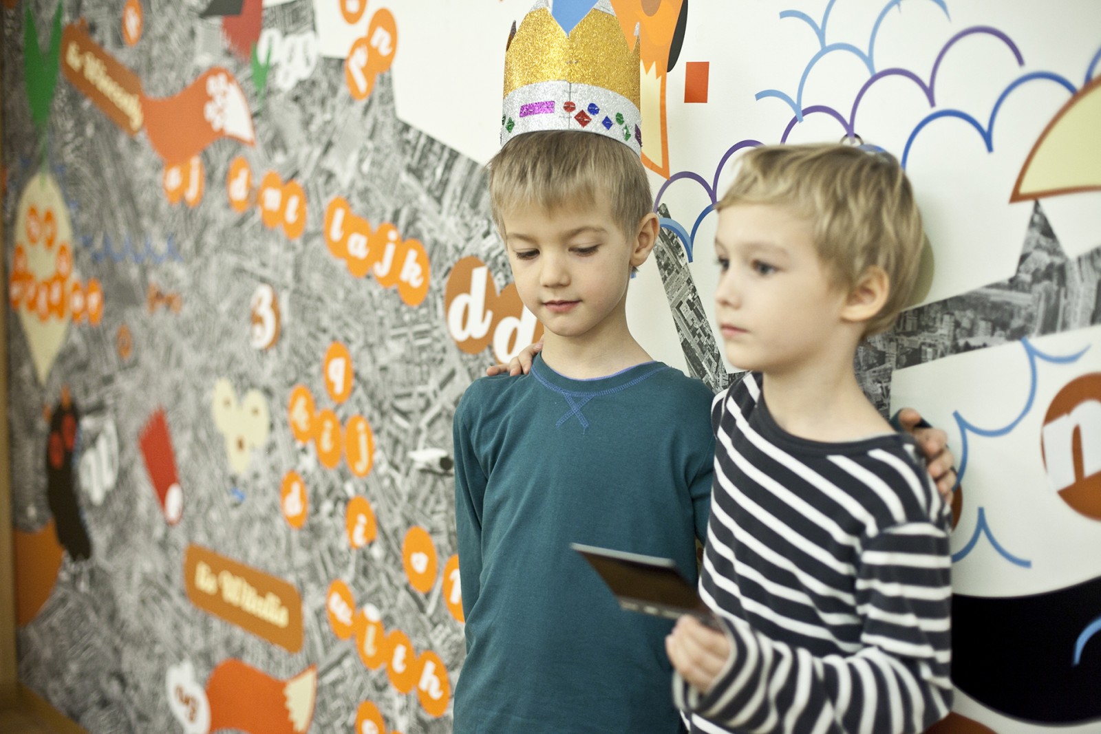 Dwóch chłopców  - po prawej stronie zdjęcia - stoi obok siebie, opierają się plecami o ścianę. Rozmawiają. Chłopiec po lewej ma na głowie kolorowa koronę.