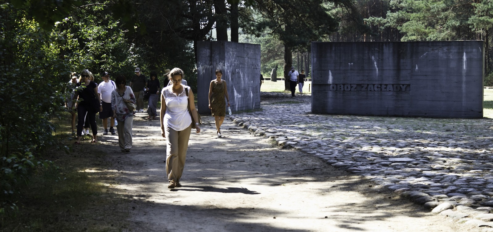 Kilka osób spaceruje w pobliżu tablicy z napisem obóz Zagłady.