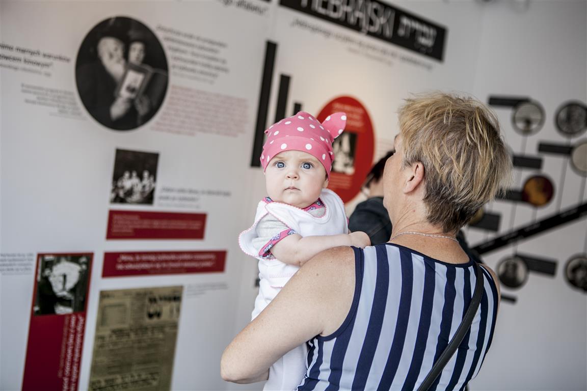 Kobieta w pasiastej bluzce oglądająca fragment mobilnej wystawy trzyma na rękach niemowlę w kolorowym ubraniu, które czymś zaciekawione patrzy w stronę obiektywu.