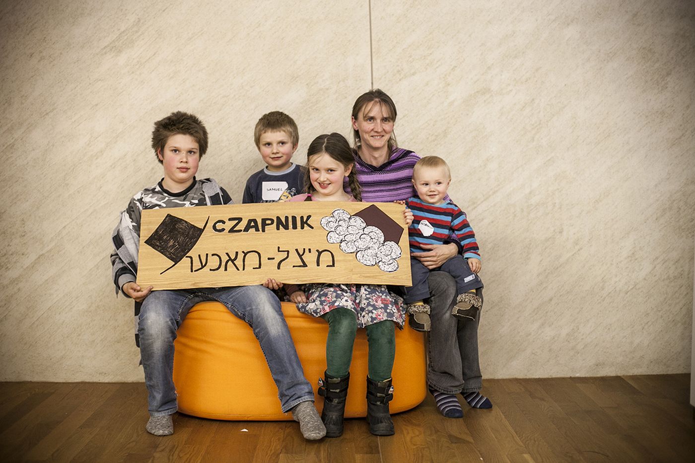 Kilkoro dzieci siedzi na pufie. Dwoje z nich trzyma tabliczkę z napisem Czapnik.