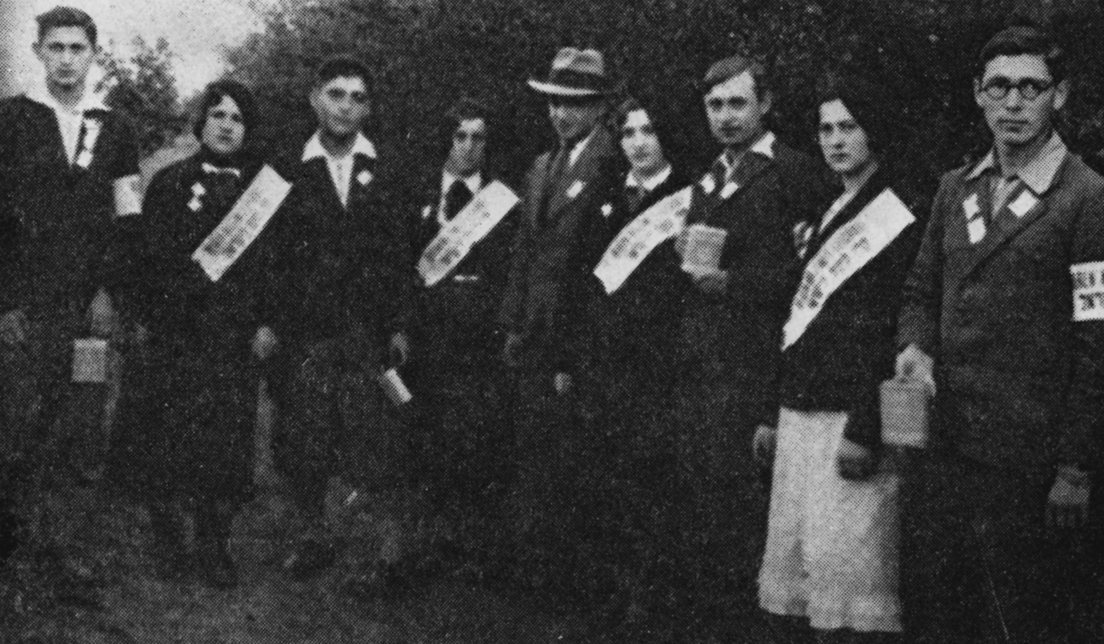Czarno-białe zdjęcie. Grupa elegancko ubranych kobiet i mężczyzn pozuje do zdjęcia. Kobiety przepasane są białymi szarfami.
