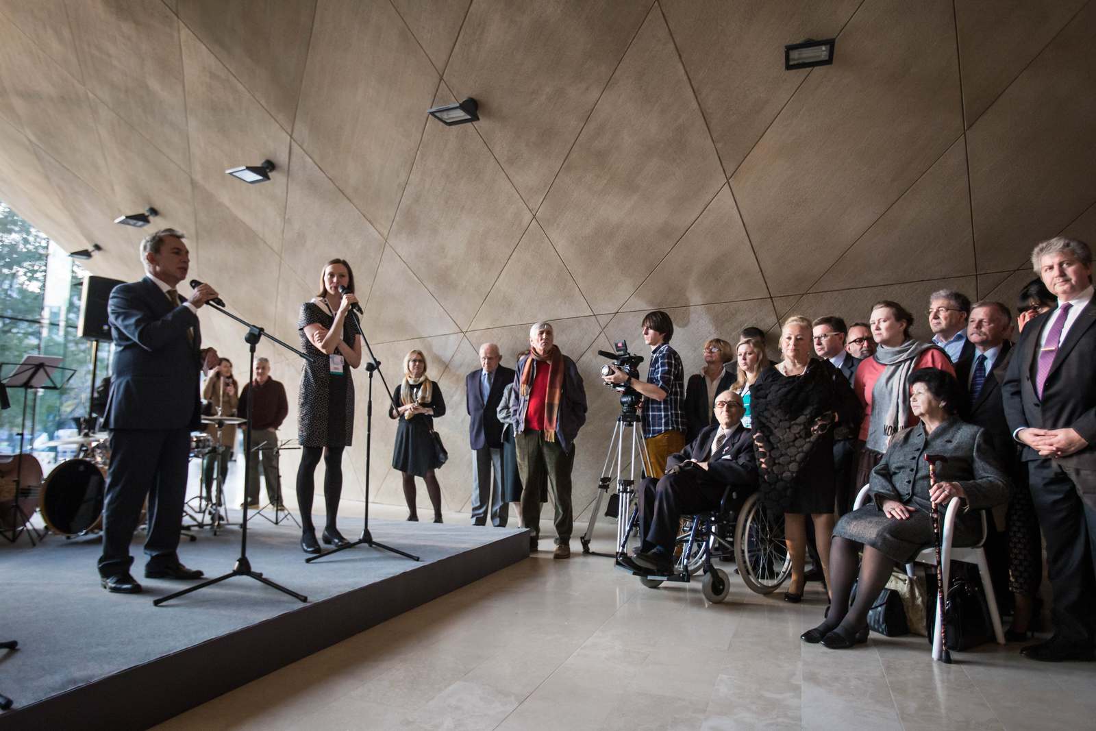 Goście w holu Muzeum POLIN słuchają mężczyzny przemawiającego ze sceny.