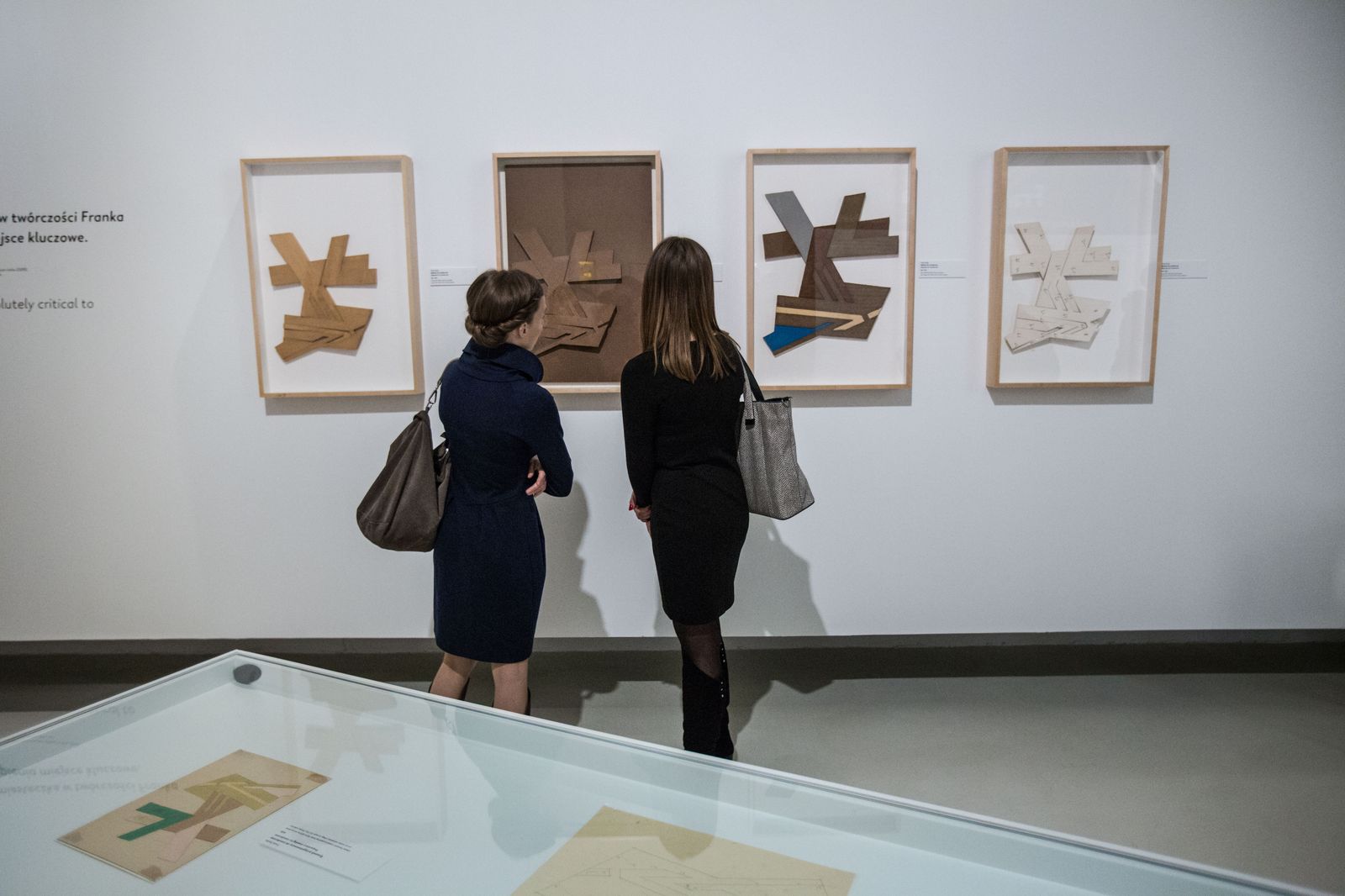 Sala w muzeum. Dwie kobiety oglądają obrazy na wystawie „Frank Stella i synagogi dawnej Polski”. Przed nimi na ścianie wiszą cztery obrazy.  