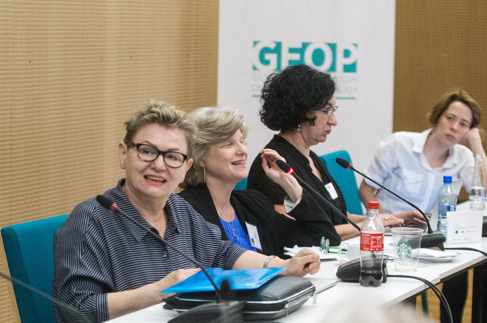 Warsztaty GEOP - cztery kobiety siedzą przy stole konferencyjnym, przed nimi mikrofony, notatki i skrypty, Kobieta pierwsza od lewej jest zwrócona twarzą do oglądających i mówi do osób poza kadrem zdjęcia.