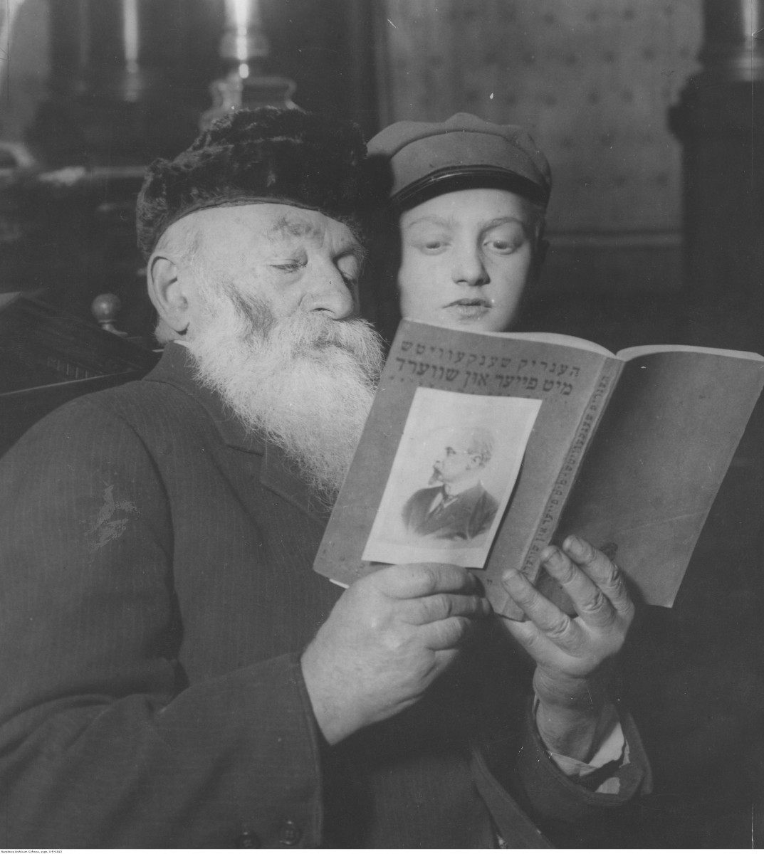 Stary Żyd z długą brodą czyta książkę zza ramienia zagląda mu w nią młody chłopiec w czapce