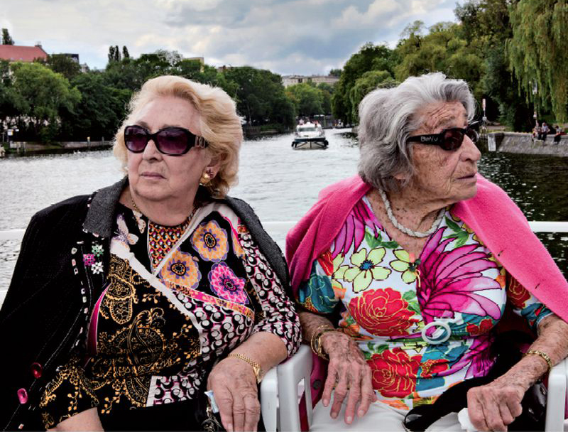 Oma & Bella - film, TISZ Festiwal Żydowskiego Jedzenia - kadr z filmu, przedstawia dwie starsze kobiety, które siedzą obok siebie z głowami odwróconymi od siebie. Każda ma ciemne okulary, narzucony na ramiona sweter oraz barwne bluzki. W tle widok rzeki i miasta położonego na jej brzegach.