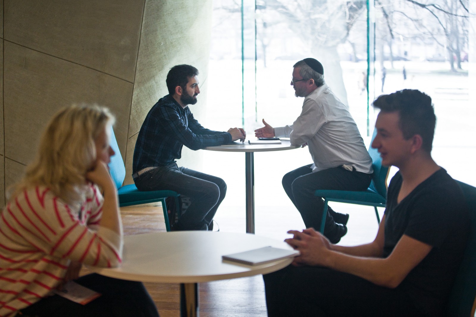 Dwie pary osób rozmawiają ze sobą przy dwóch oddzielnych stolikach w przestrzeni kawiarni Muzeum Polin
