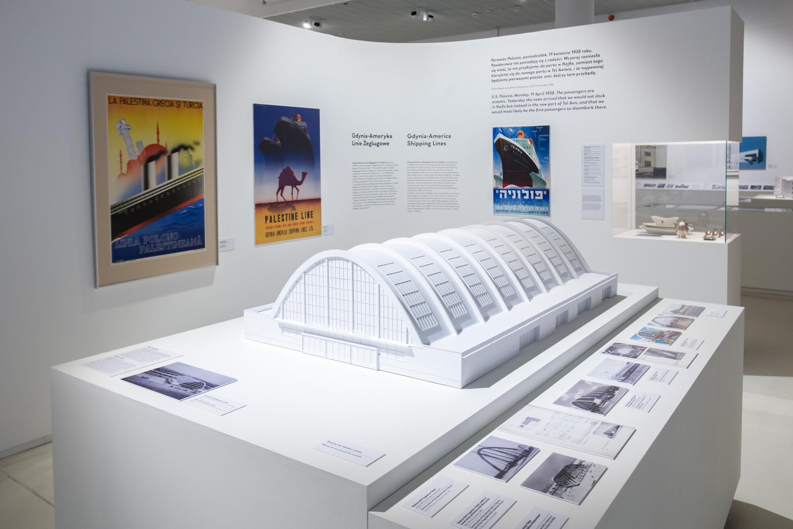 Wystawa czasowa "Gdynia - Tel Awiw" w Muzeum POLIN. Na zdjęciu biały model architektoniczny hali targowej w Gdyni, widać żebrowanie półokrągłych elementów konstrukcyjnych i inne składowe bryły. W tle, na białej ścianie obok, plakaty prezentowany na wystawie