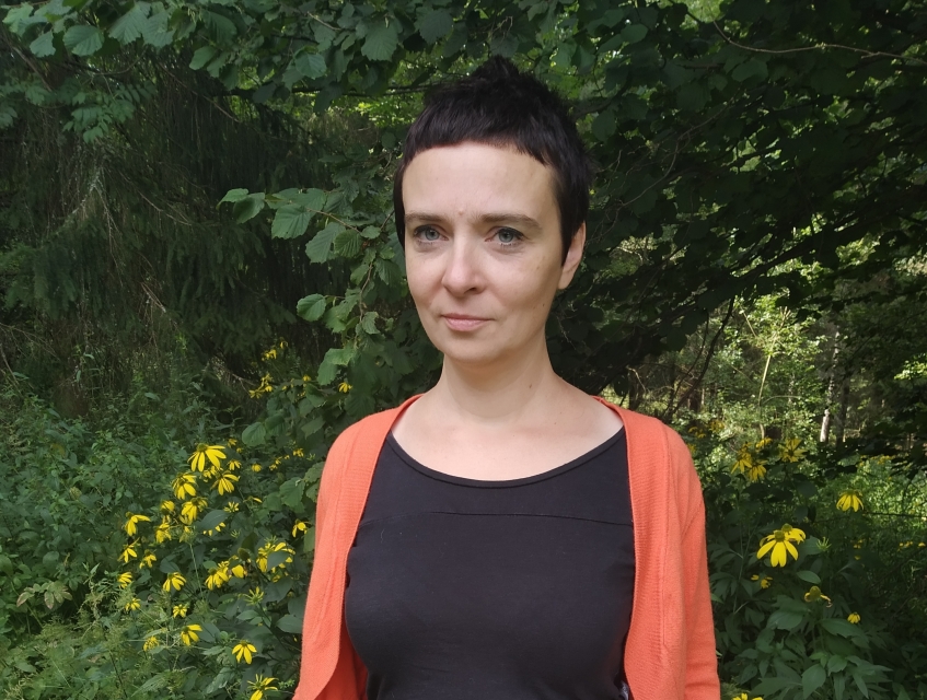Na zdjęciu widać kobietę na tle zieleni roślin. Ma krótkie ciemne włosy. To Patrycja Dołowy