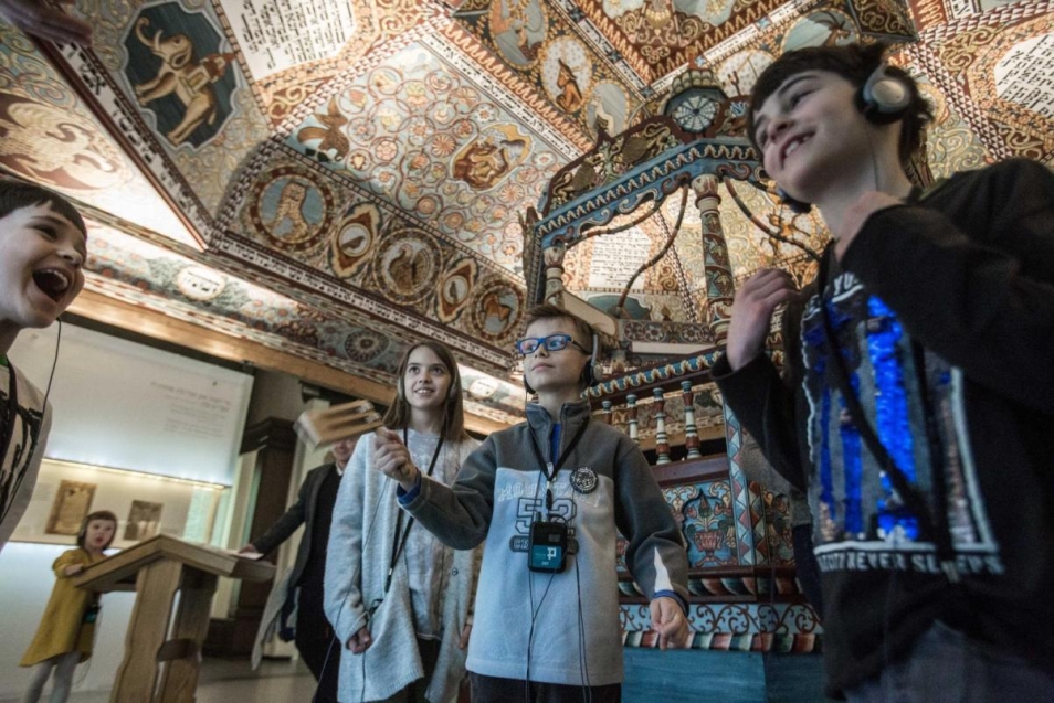 Grupa dzieci znajduje się na wystawie Muzeum POLIN pod dachem rekonstrukcji synagogi w Gwoźdźcu. Dzieci mają na uszach słuchawki od audioprzewodników.
