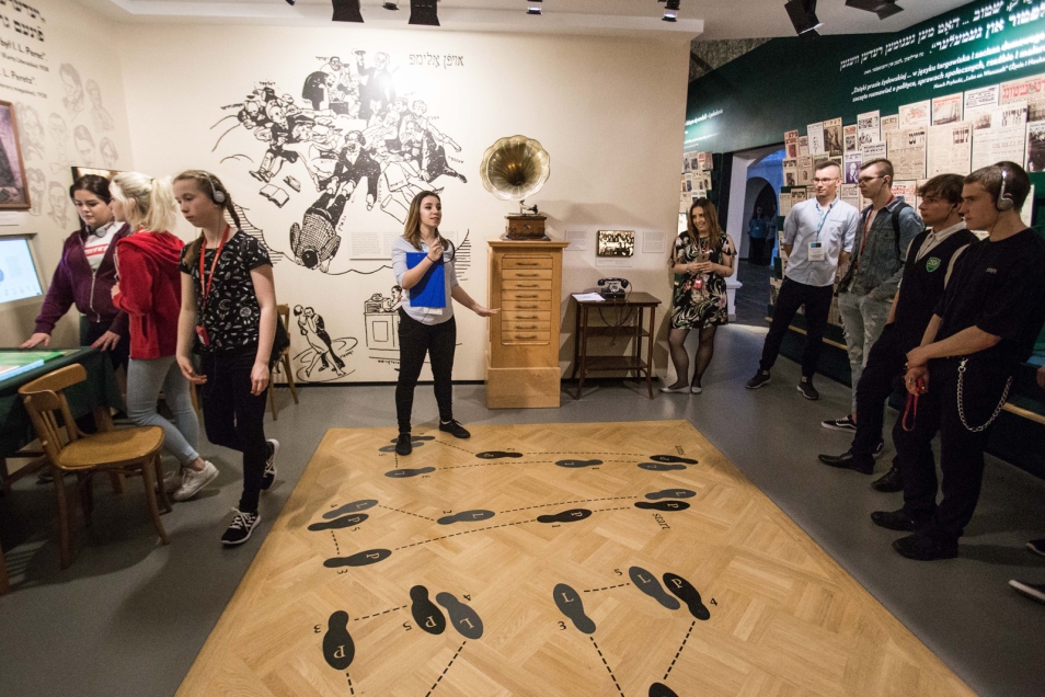 Na zdjęciu grupa młodzieży znajduje się w galerii Miasteczko na wystawie stałej Muzeum POLIN, w sali tanecznej, próbują tańczyć tango wg kroków rozrysowanych na podłodze