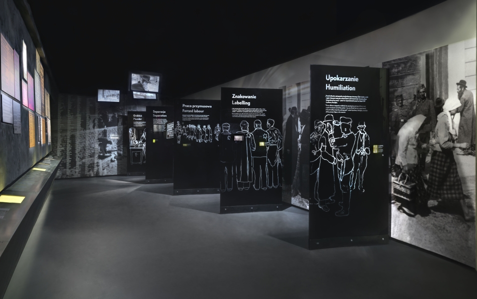Widok na fragment galerii Zagłada na wystawie Muzeum POLIN. Wnętrze w kolorach czarnym i ciemnym szarym. Przedstawione są wnim, na czarnych ściankach, zdjęcia i opisy sposobu prześladowania Żydów przez żołnierzy nazistowskich.