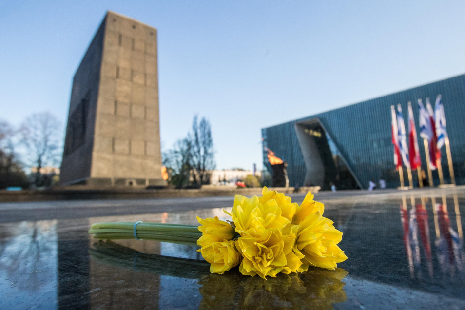 Na wypolerowanej kamiennej, ciemnej płycie leży wiązanka żonkili. W tle widać zarys pomnika Bohaterów Getta w Warszawie i budynek Muzeum POLIN