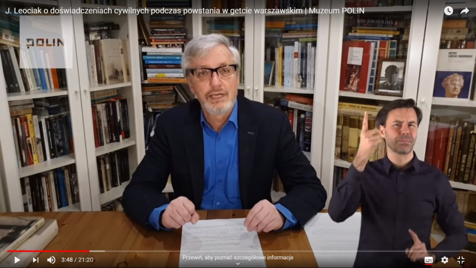 Na zdjęciu, przy biurku siedzi mężczyzna w okularach - prof. Jacek Leociak - w prawym dolnym rogu widać tłumacza PJM