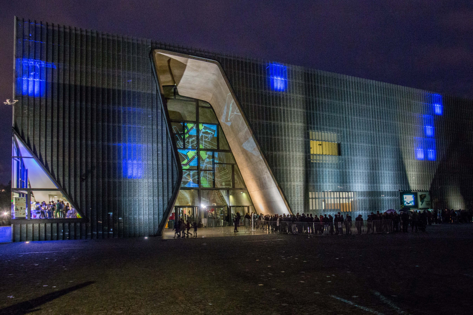 Oświetlony na kolorowo budynek Muzeum POLIN. Przed wejściem kolejka ludzi.