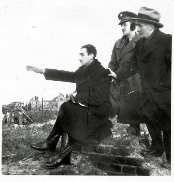 Czarno-białe historyczne zdjęcie. Trzech mężczyzn. Jeden siedzi na murze (Marek Edelman) i wskazuje wyciągniętą ręką w lewą stronę. Za nim dwóch mężczyzn patrzy w tę samą stronę. Jeden trzyma siedzącego za ramię. W tle ruiny miasta.