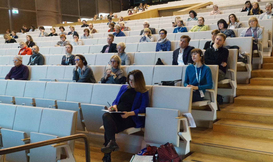 Sala konferencyjna - widok na ustawione w rzędach fotele. W niektórych siedzą uczestnicy konferencji. Słuchają i notują